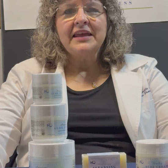 Woman in a lab coat presents a variety of skincare products on a table, including creams and cleansers.