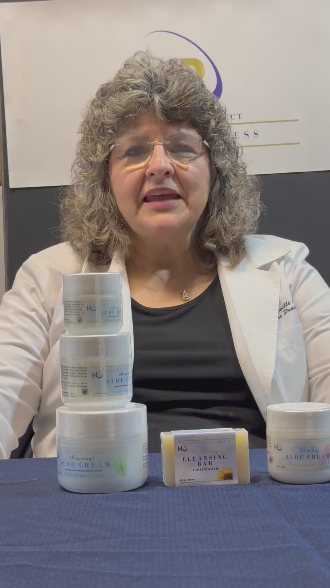 Woman in a lab coat presents a variety of skincare products on a table, including creams and cleansers.